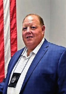 Undersheriff Cole in blue suit coat with white shirt and badge hanging from neck.